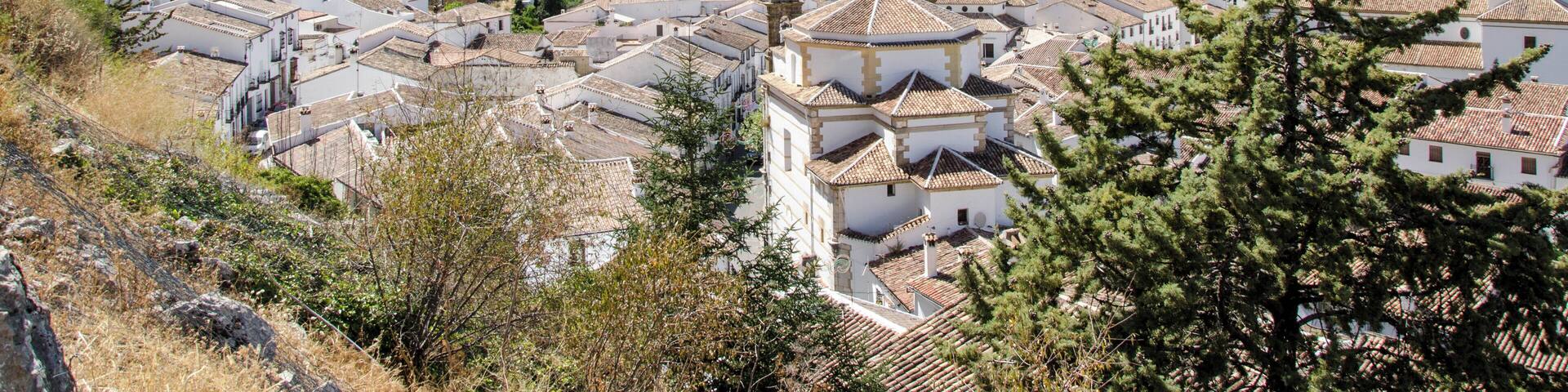 A mountainous town with white buildings and terracotta roofs, surrounded by lush green hills and a prominent rocky peak.