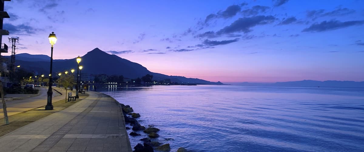 A waterfront promenade at dusk with street lamps and a mountain in the background.