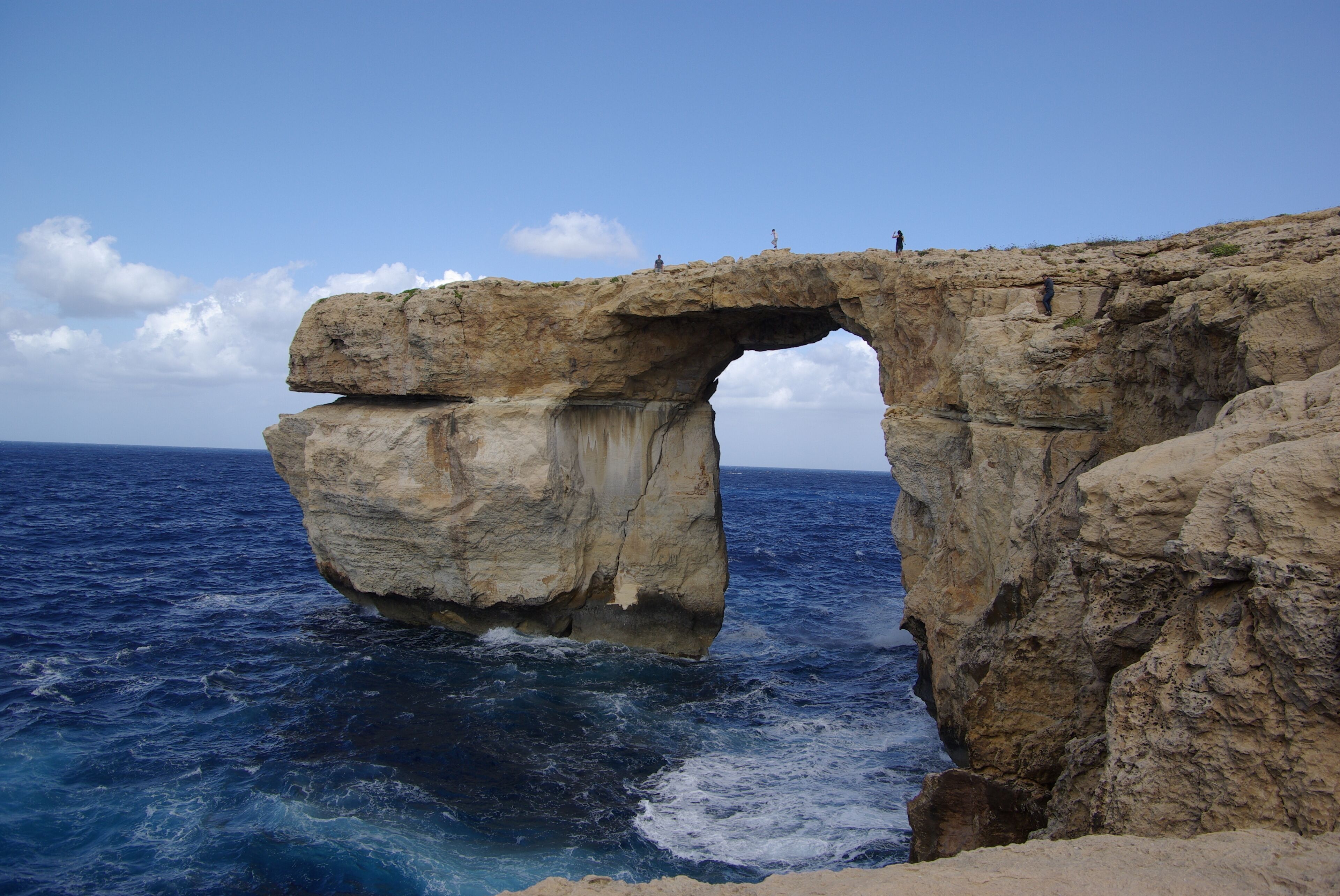 A large rock arch over the sea with people standing on top.