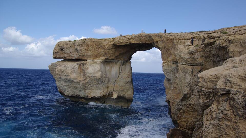 A large rock arch over the sea with people standing on top.