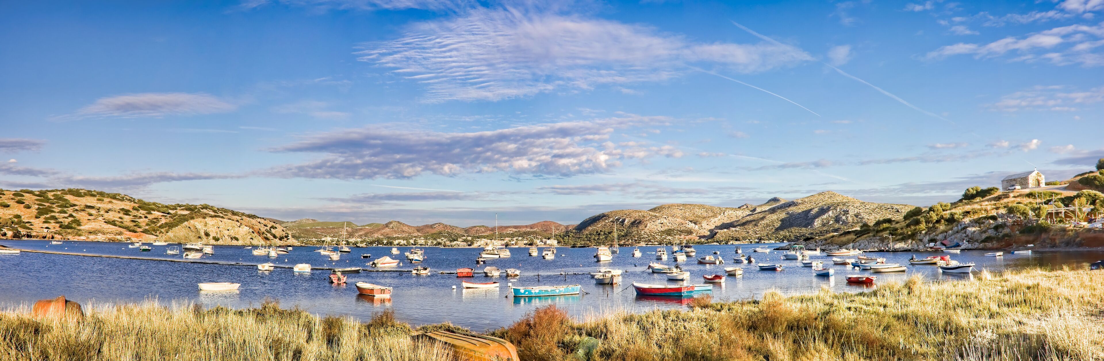 A coastal bay with numerous boats anchored in calm waters, surrounded by rocky hills and sparse vegetation.