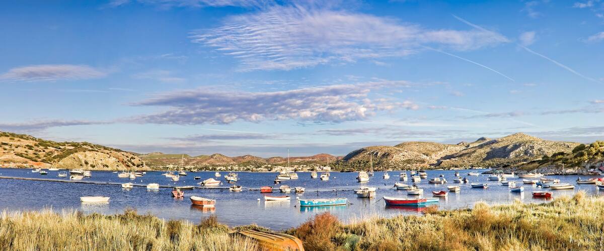 A coastal bay with numerous boats anchored in calm waters, surrounded by rocky hills and sparse vegetation.