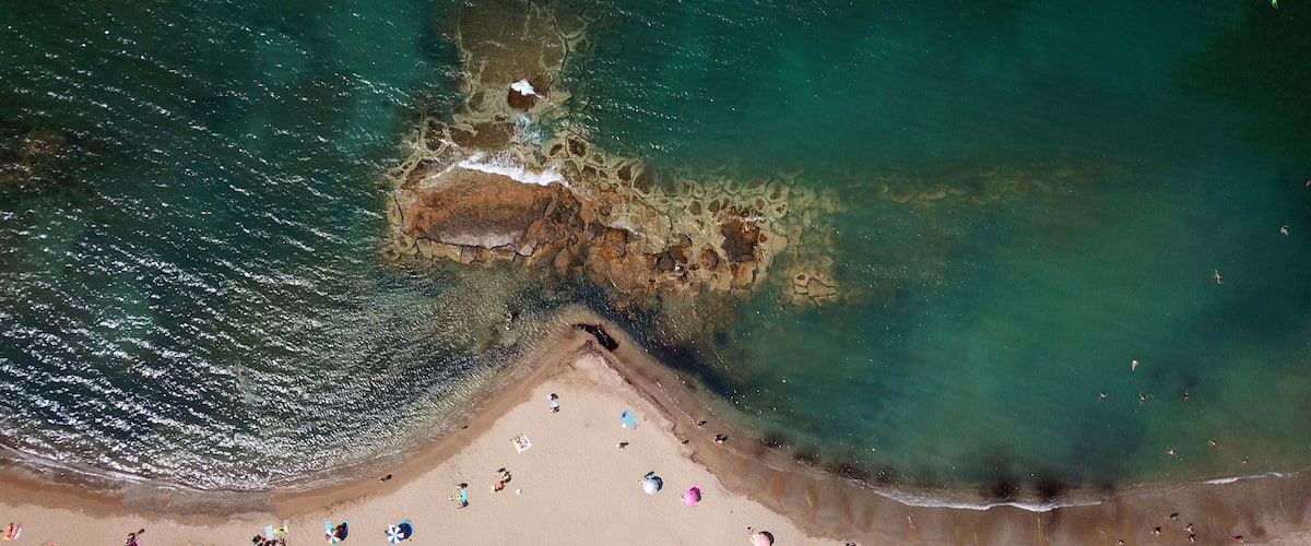 Vista aérea de una playa con aguas turquesas cristalinas, un afloramiento rocoso y gente tomando el sol.