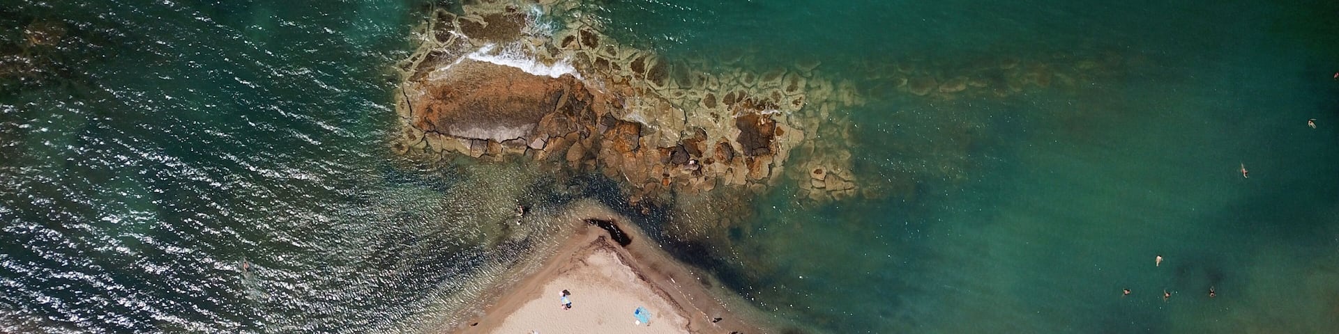 An aerial view of a beach with clear turquoise waters, a rocky outcrop, and people sunbathing.