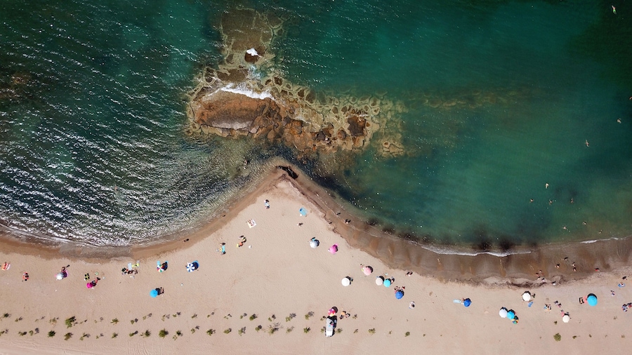 An aerial view of a beach with clear turquoise waters, a rocky outcrop, and people sunbathing.
