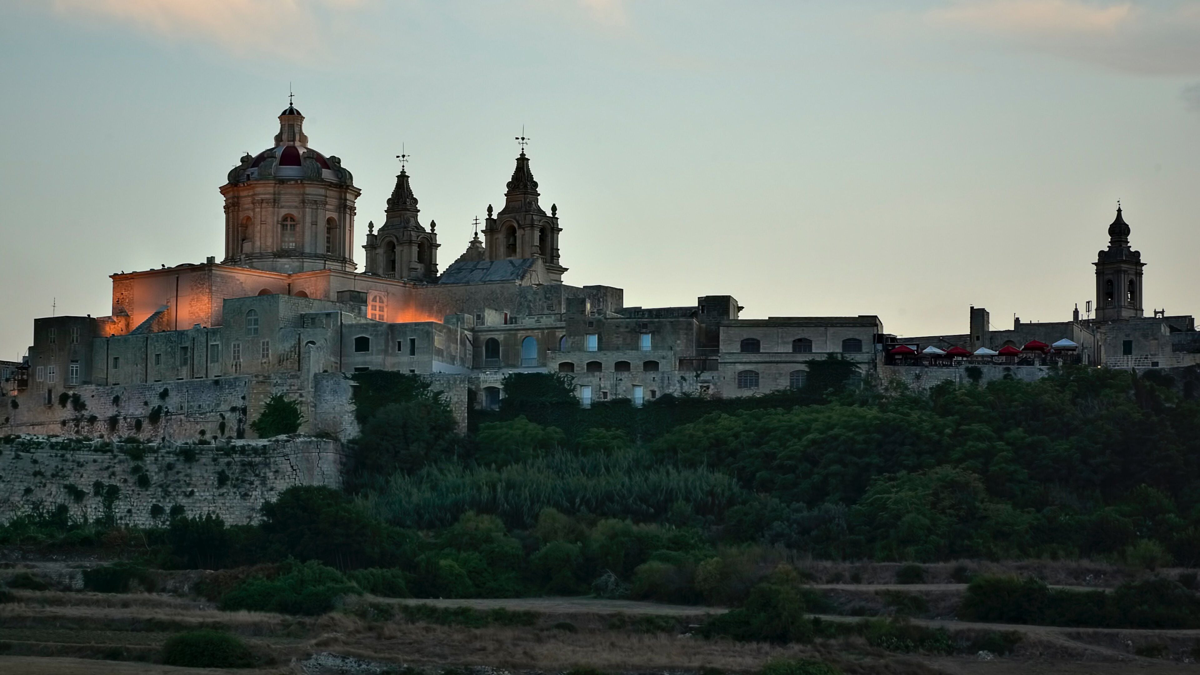 A historic cityscape with a prominent cathedral featuring multiple domes and spires, surrounded by lush greenery and terraced fields.