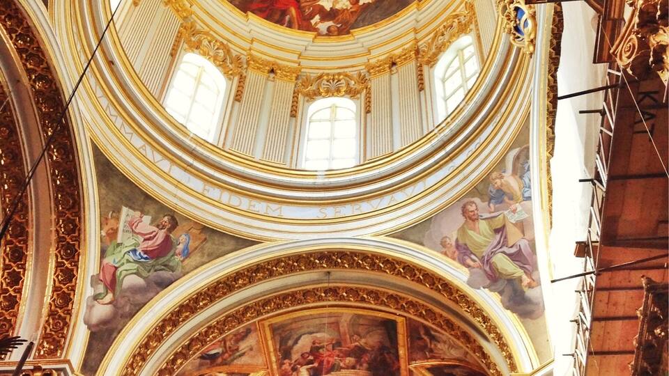 A grand, ornate church interior with a high, circular ceiling, intricate frescoes, and a central stained glass window.