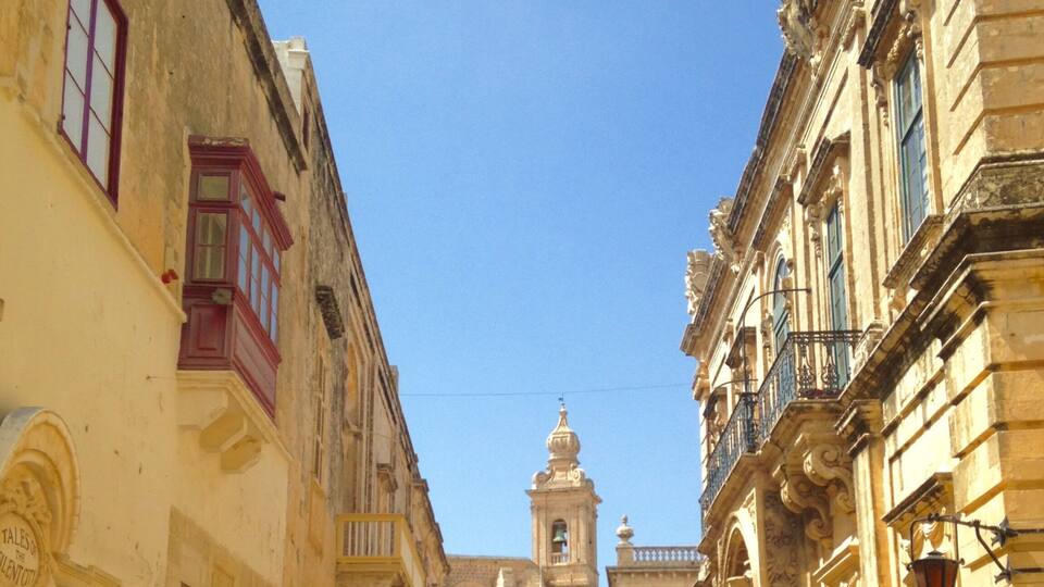 A narrow street with historic buildings, a bell tower, and a clear blue sky.