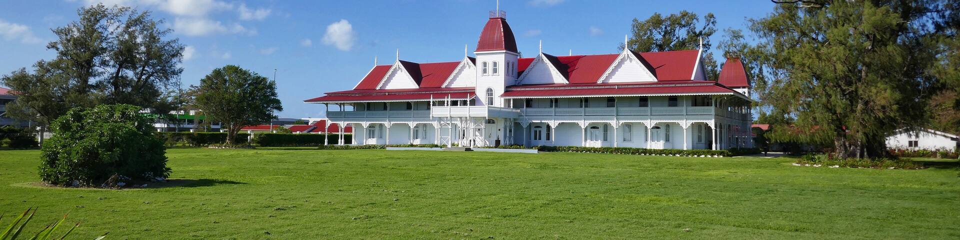 A large white building with a red roof and multiple chimneys, surrounded by a well-maintained lawn and trees.