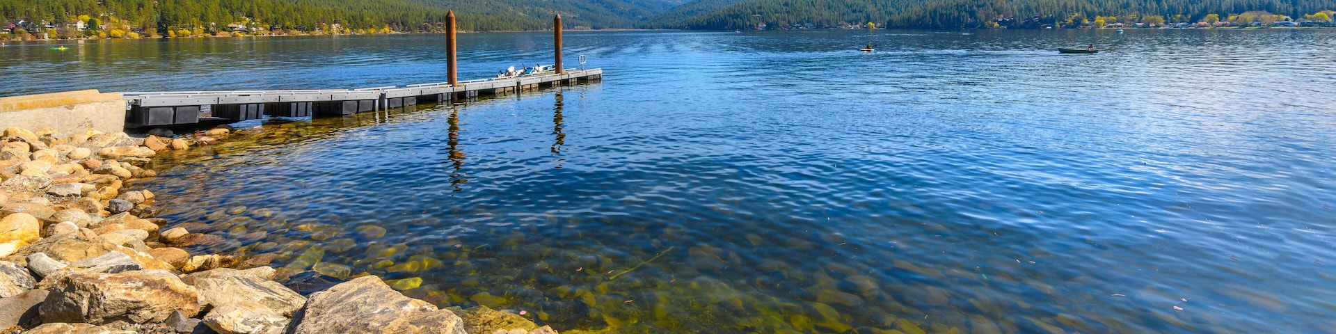 Un lago tranquilo con aguas cristalinas, una orilla rocosa, un muelle de madera y colinas boscosas al fondo.