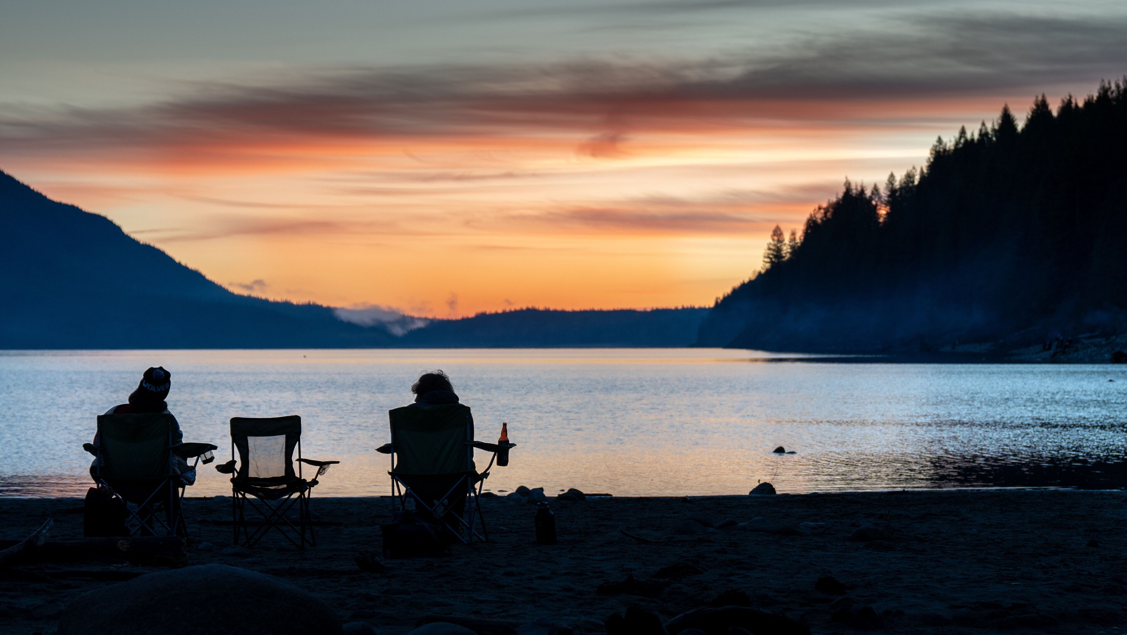 A sunset over a calm lake with two people sitting on chairs facing the water, surrounded by mountains and trees.