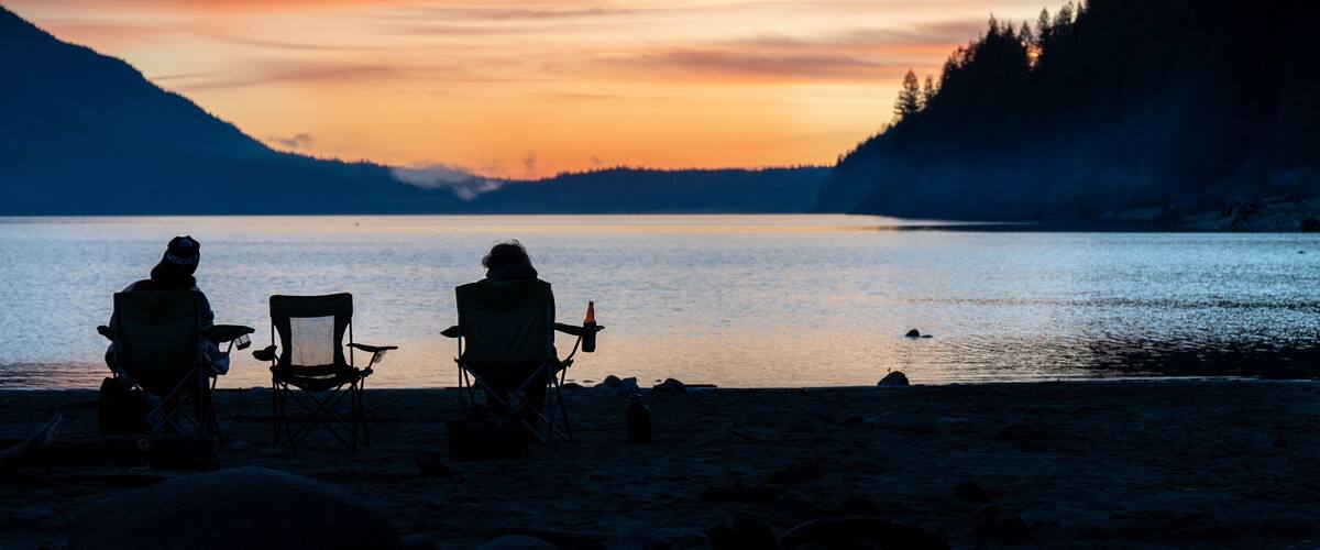 A sunset over a calm lake with two people sitting on chairs facing the water, surrounded by mountains and trees.