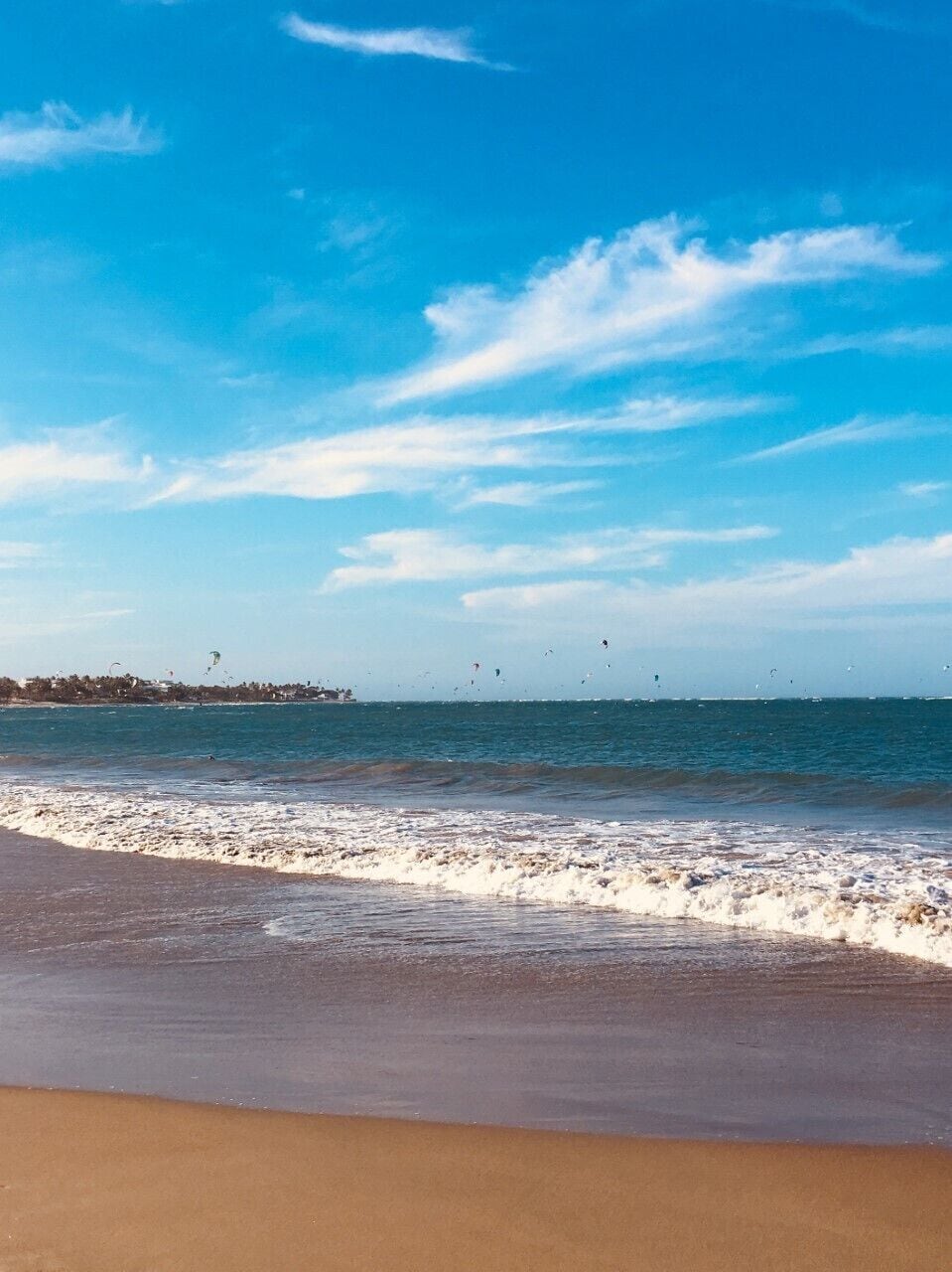 A beach with waves, a clear sky, and kites flying.