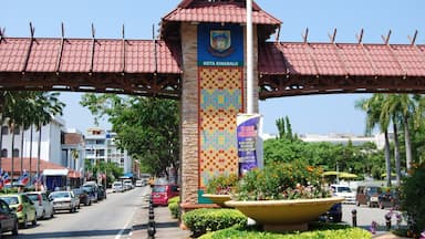 Uma rua com uma placa de entrada destacada para Kota Kinabalu, rodeada por palmeiras e carros estacionados.