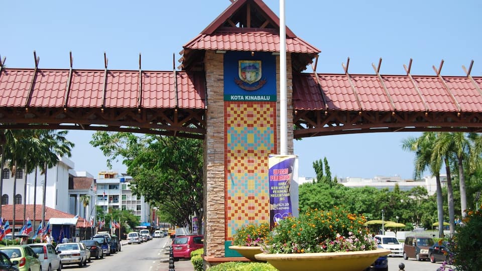 A street with a prominent entrance sign for Kota Kinabalu, surrounded by palm trees and parked cars.