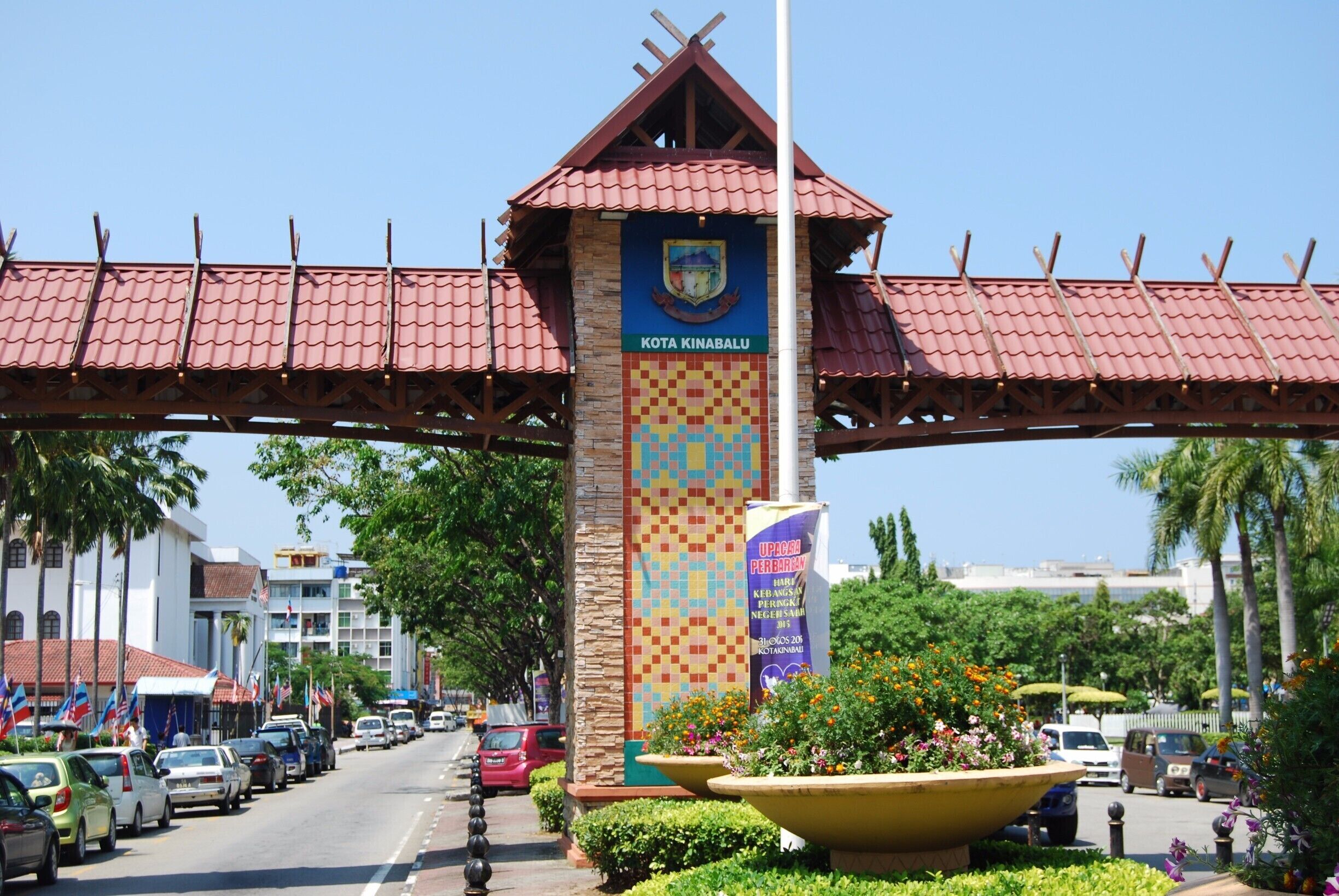 A street with a prominent entrance sign for Kota Kinabalu, surrounded by palm trees and parked cars.