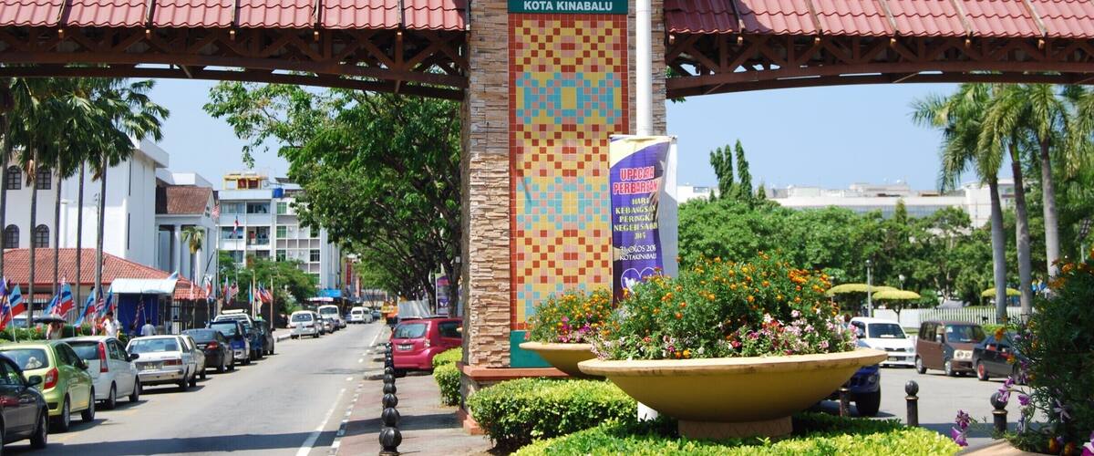 A street with a prominent entrance sign for Kota Kinabalu, surrounded by palm trees and parked cars.