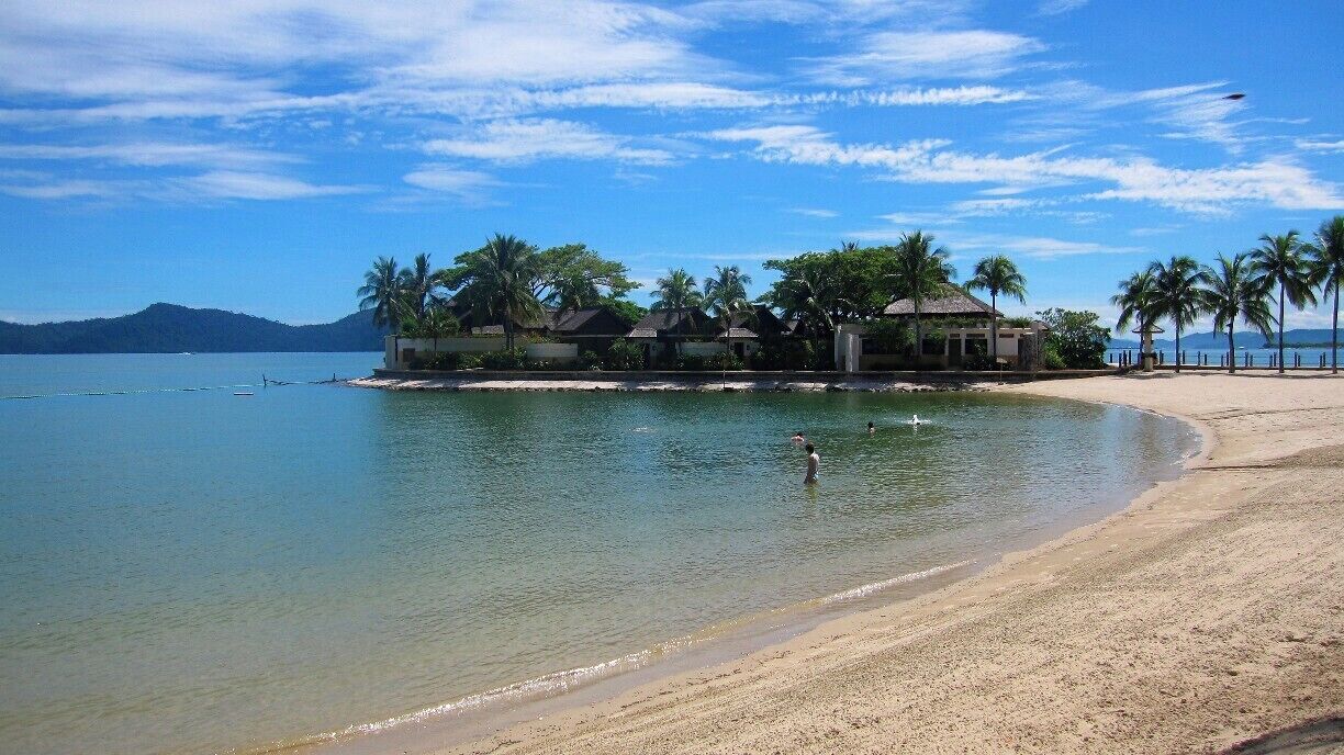 A tropical beach with clear water, palm trees, and a resort in the background.