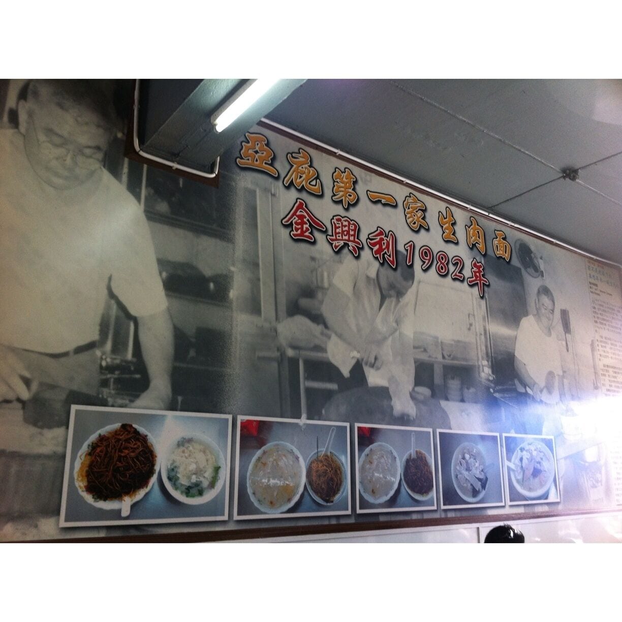 Barraca de comida com um letreiro mostrando diversos pratos e uma placa indicando que o estabelecimento está em funcionamento desde 1982.