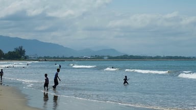 Uma praia com pessoas brincando na água e um avião na pista, ao fundo.