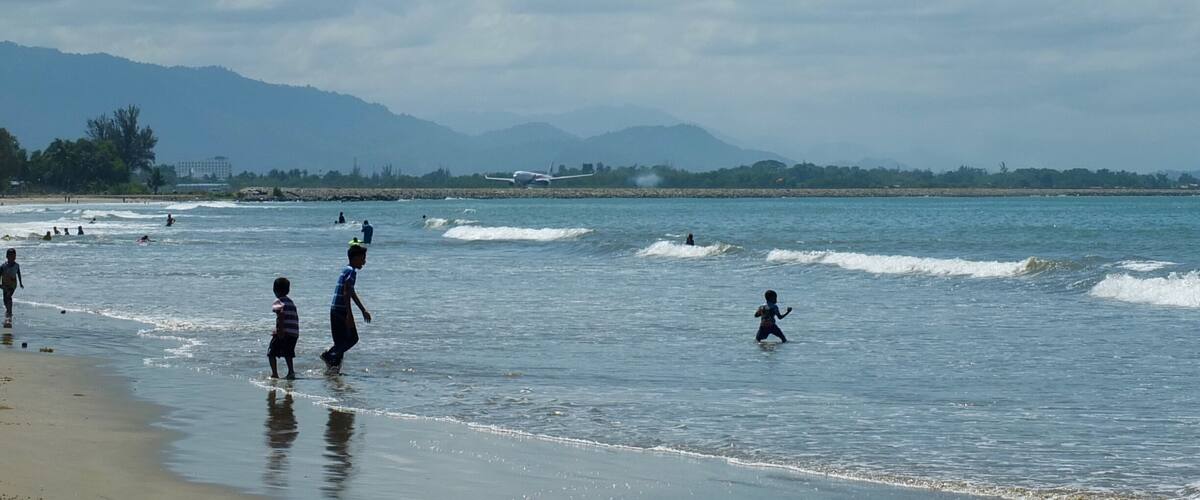 A beach with people playing in the water and a plane on the runway in the background.