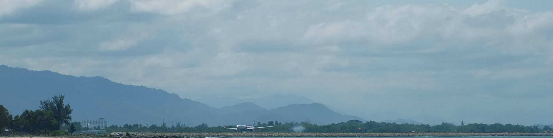 A beach with people playing in the water and a plane on the runway in the background.
