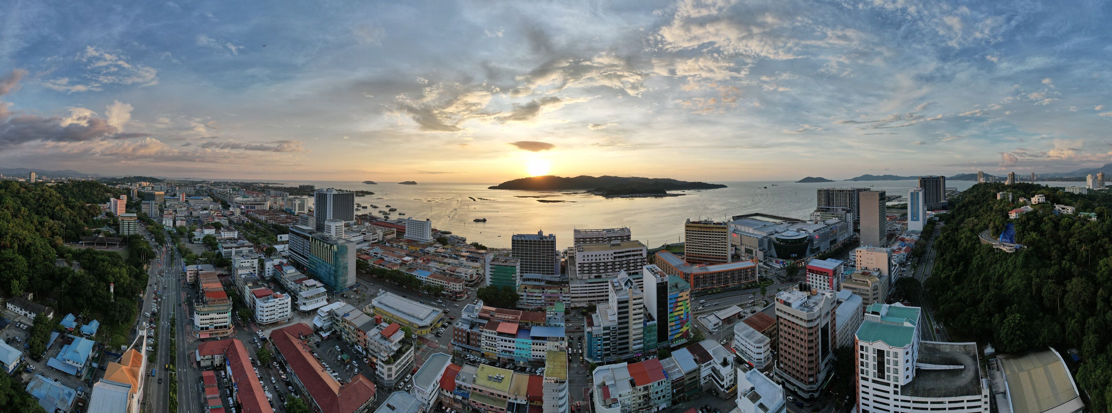 A coastal cityscape with a sunset over the sea, featuring dense urban buildings and a clear sky.
