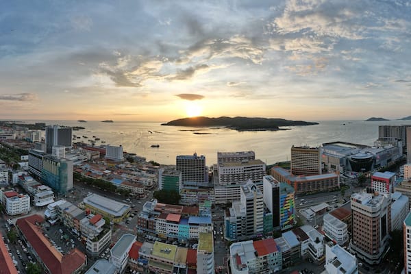 A coastal cityscape with a sunset over the sea, featuring dense urban buildings and a clear sky.