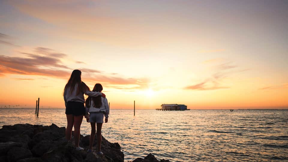 A sunset over the ocean with a pier and a house on stilts in the distance.