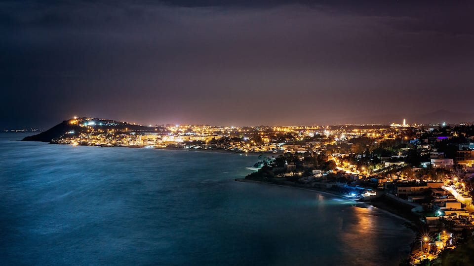 A coastal city at night with a prominent hilltop and a curved coastline.
