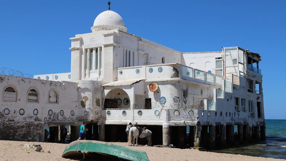 A white building with a dome and arched windows, situated on a sandy beach with the ocean in the background.