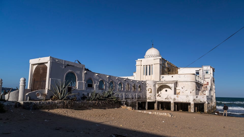 A coastal building with a dome and arched windows, adjacent to a sandy beach with the ocean in the background.