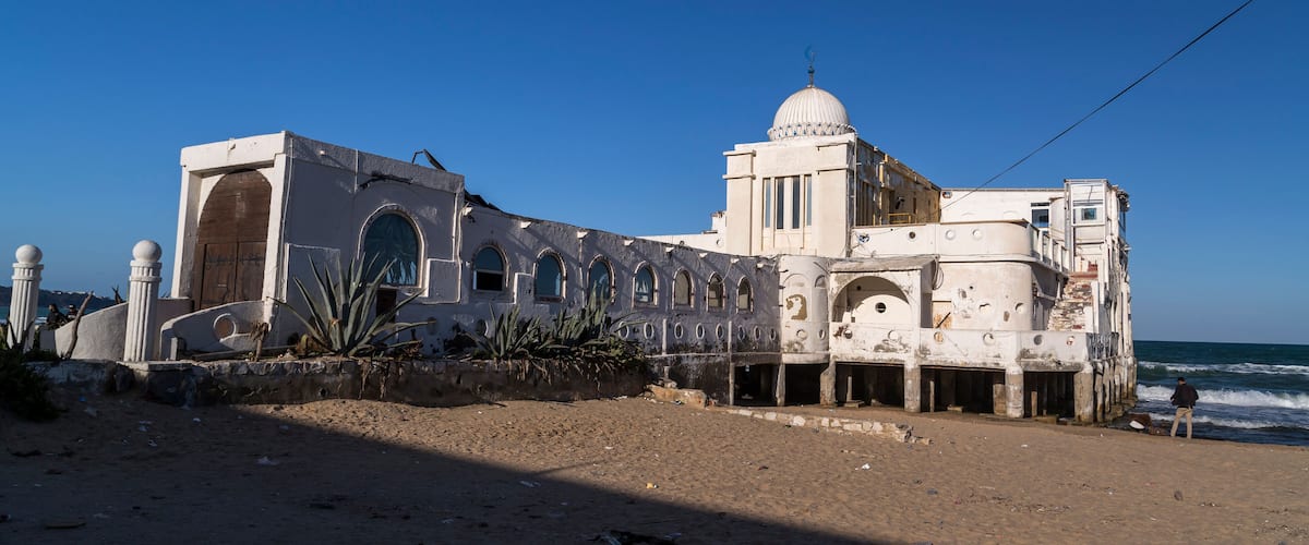 A coastal building with a dome and arched windows, adjacent to a sandy beach with the ocean in the background.