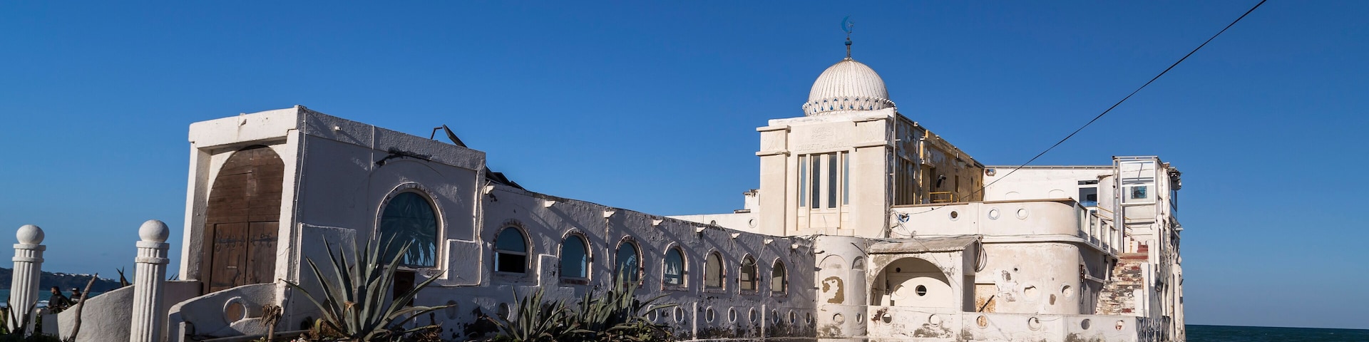A coastal building with a dome and arched windows, adjacent to a sandy beach with the ocean in the background.