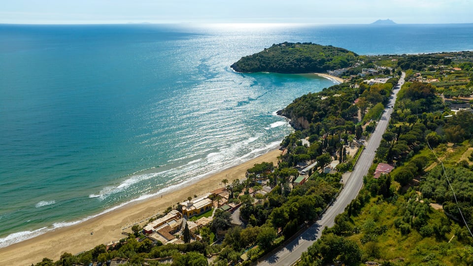 A coastal landscape with a beach, a road, and a hill with vegetation.