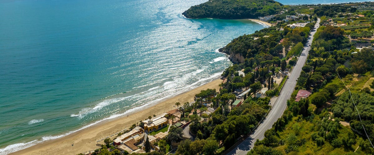 A coastal landscape with a beach, a road, and a hill with vegetation.