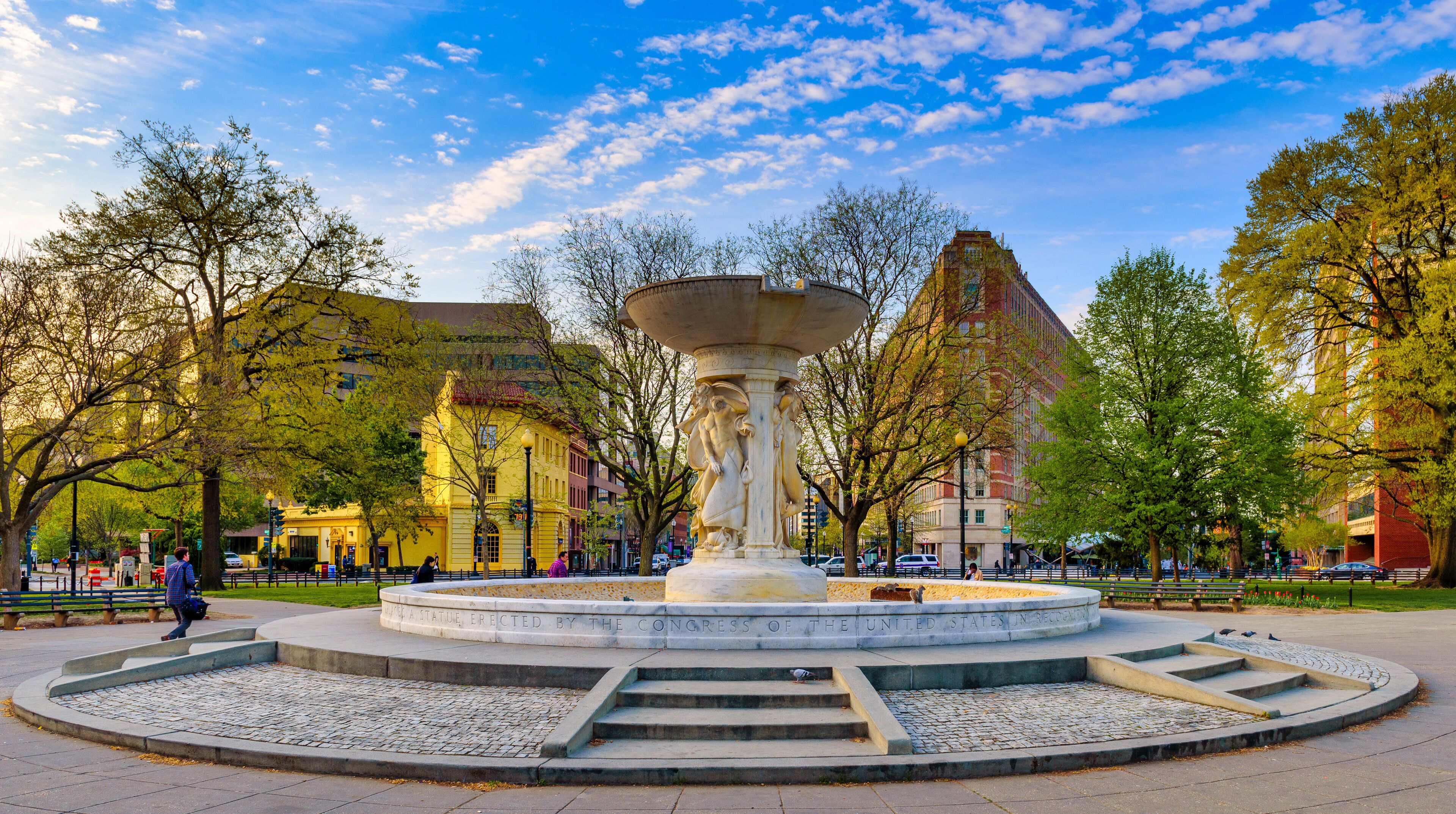 A park with a central fountain, surrounded by trees and buildings.