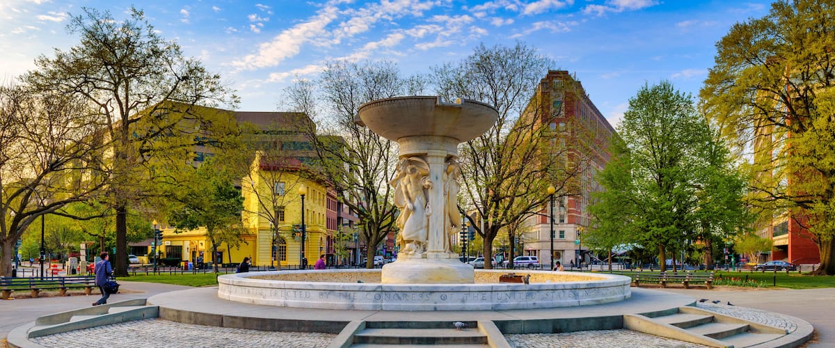 A park with a central fountain, surrounded by trees and buildings.