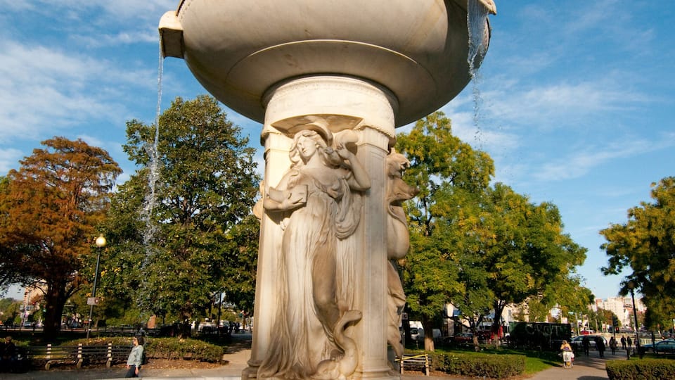 A classical fountain with a large basin and sculpted figures, surrounded by a park with trees and a clear sky.