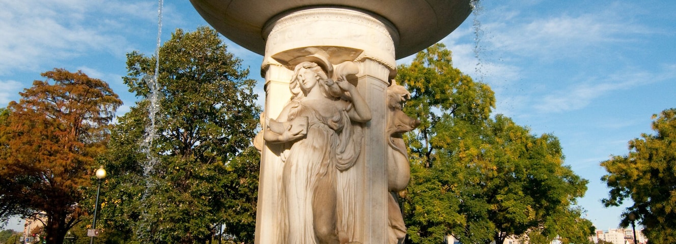 Uma fonte clássica com uma grande bacia e figuras esculpidas, rodeada por um parque com árvores e um céu limpo.