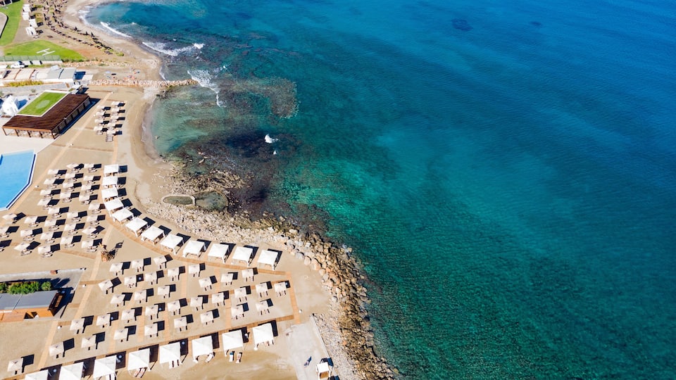 An aerial view of a beach with clear turquoise waters, a rocky shoreline, and a row of beach umbrellas and lounge chairs.
