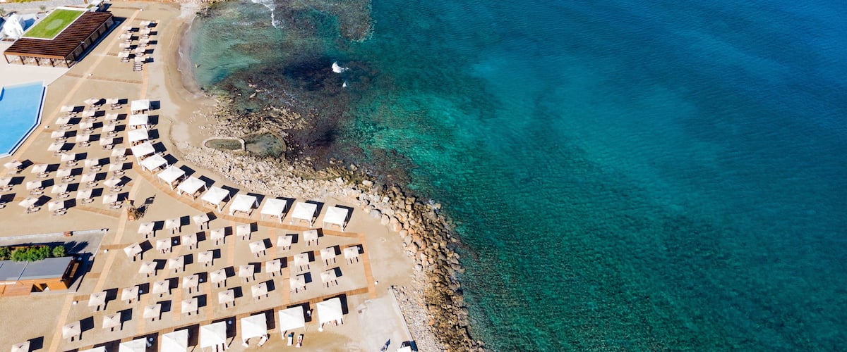 An aerial view of a beach with clear turquoise waters, a rocky shoreline, and a row of beach umbrellas and lounge chairs.