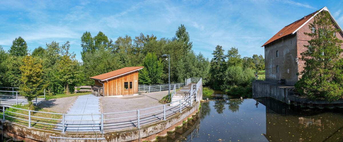 A wooden cabin and a brick building by a calm river, surrounded by trees and a clear sky.