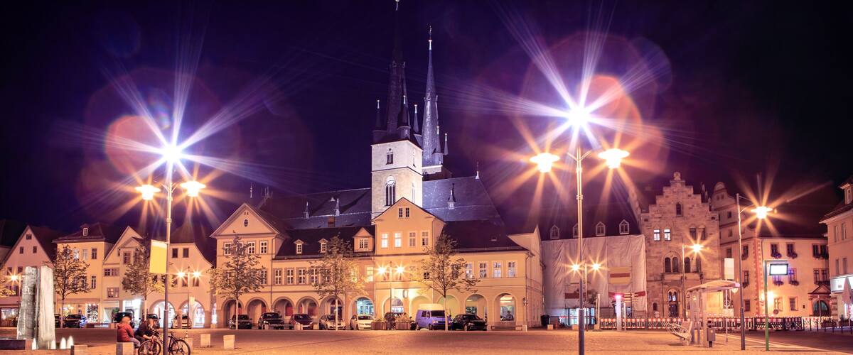 A nighttime city square with illuminated buildings, a church, and street lamps.