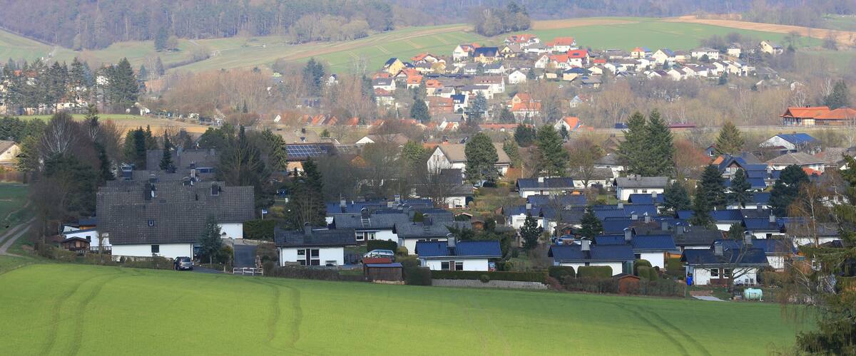 A rural village with houses, green fields, and a forested hill in the background.