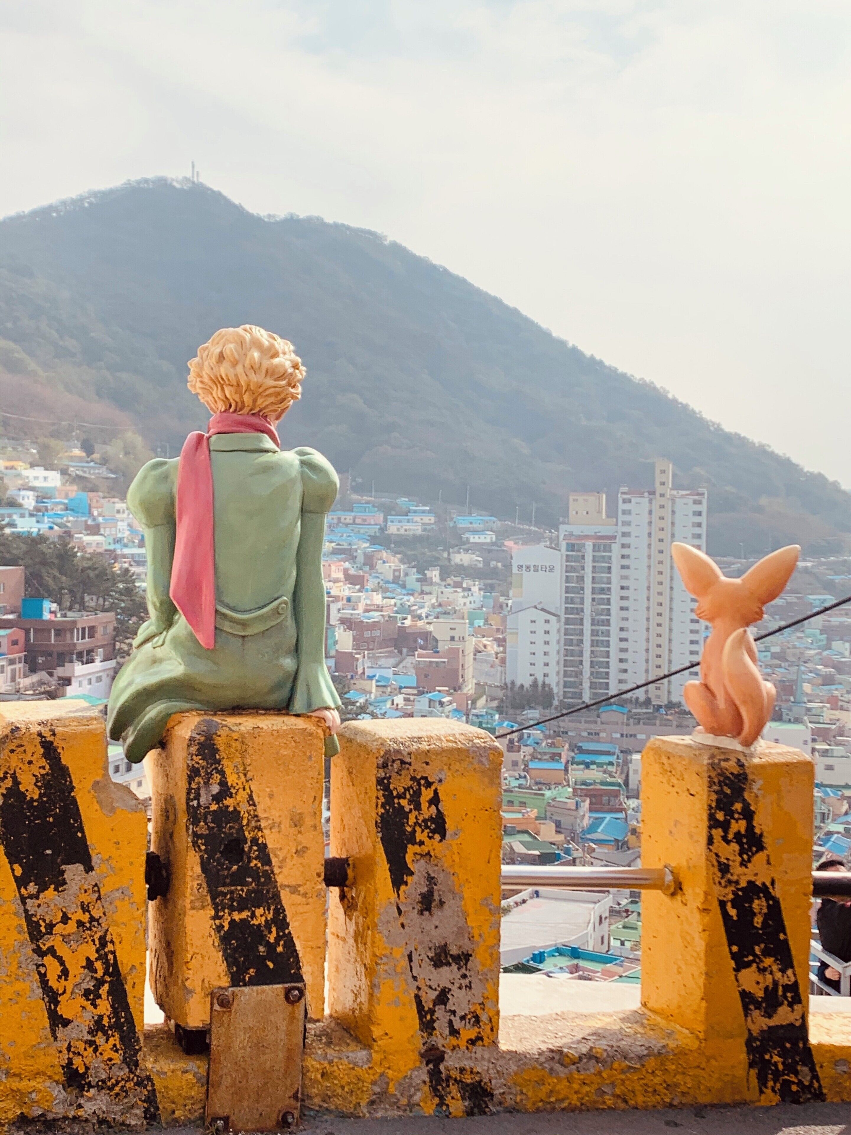 A person sitting on a yellow barrier overlooking a city with colorful buildings and a mountain in the background.