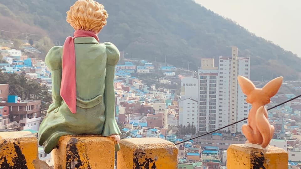 A person sitting on a yellow barrier overlooking a city with colorful buildings and a mountain in the background.