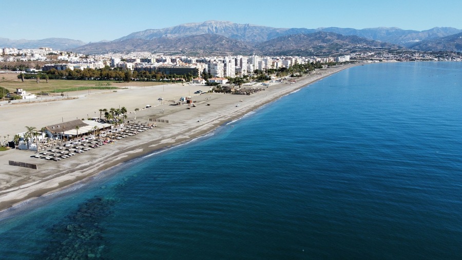 Un paisaje costero con una playa extensa, mar azul cristalino y montañas al fondo.