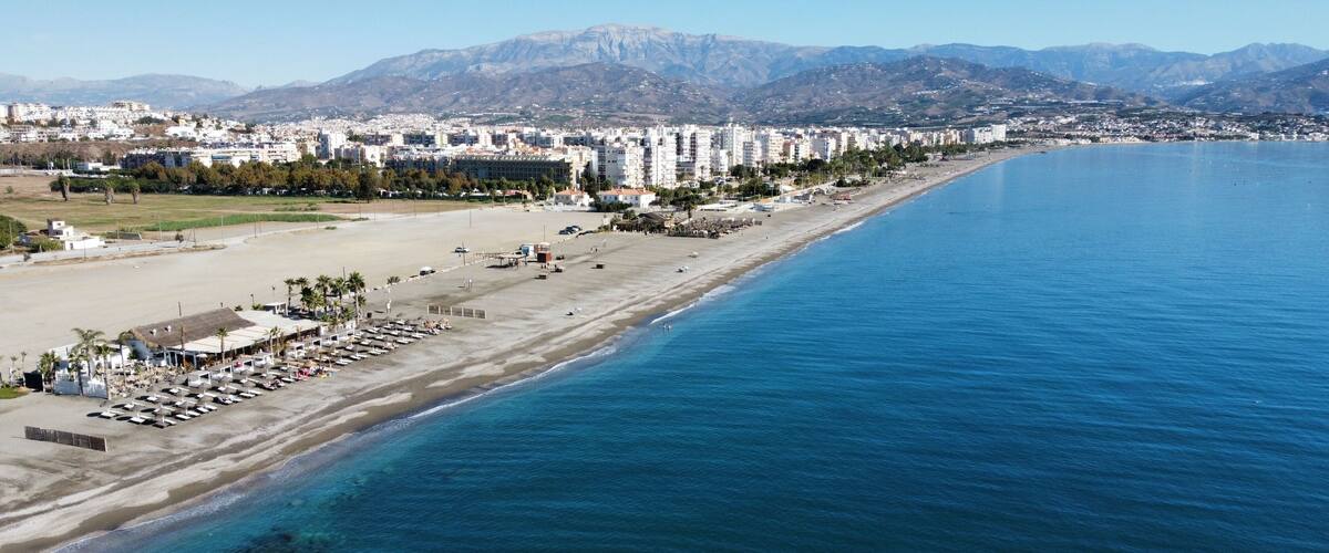 A coastal cityscape with a long beach, clear blue sea, and mountains in the background.