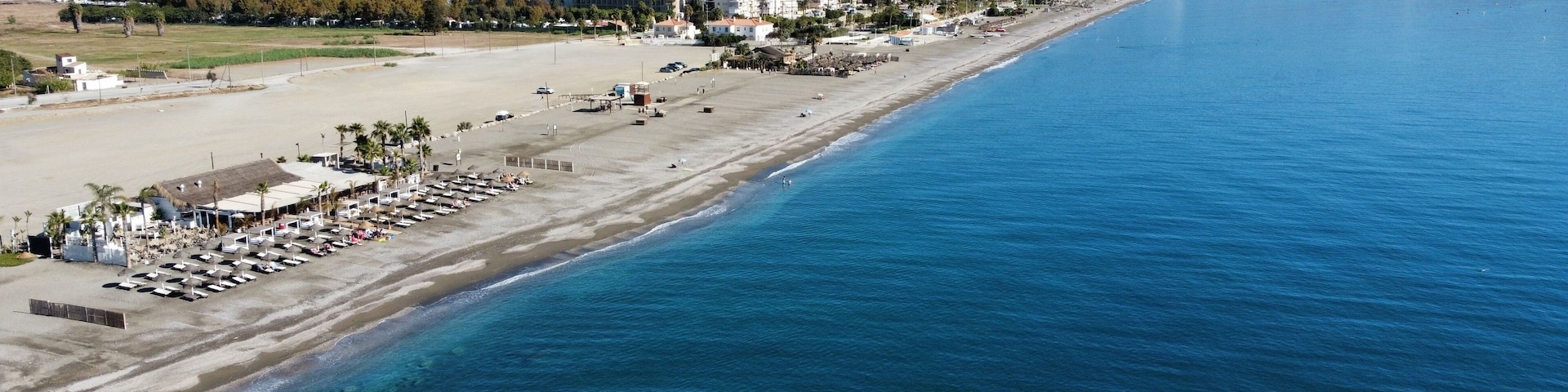 A coastal cityscape with a long beach, clear blue sea, and mountains in the background.